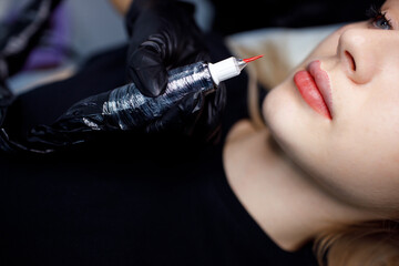 Cosmetologist applying red permanent make up tattoo on young woman lips.