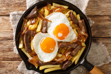Chorrillana is a dish consisting of french fries topped with sliced meat, fried eggs, and onions close up in the pan on the table. Horizontal top view from above