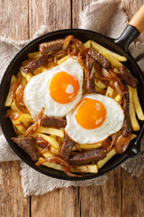 Chilean Chorrillana Fries with beef, caramelized onions, and fried eggs close up in the pan on the table. Vertical top view from above