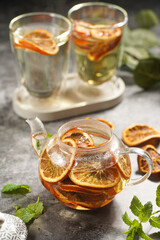 A transparent tea pot filled with black tea, oranges and mint on a grey surface