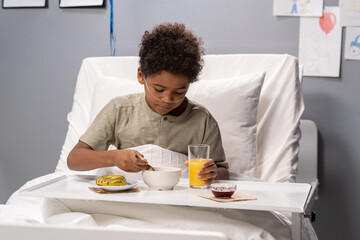 Fototapeta na wymiar Sick little boy sitting in bed with tray and having breakfast while being treated at hospital