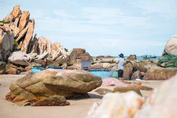 Local fisherman in the hat rolls up fish nets on the seashore. Picturesque beach with large rocks. Amazing places in the world. Real people life. 