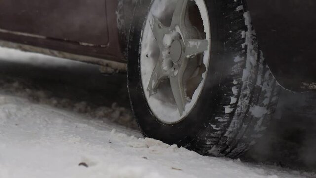 Car is standing at the snow-covered road in winter city. Smoke gases from exhaust pipe. Snowfall. Close up of wheel in slow motion. Tire. Bad weather conditions for traffic, blizzard. Danger for trip