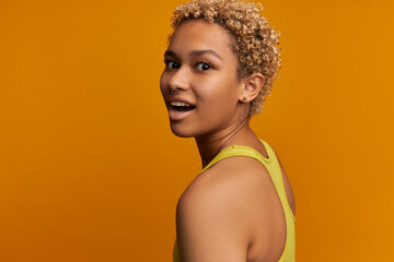 Half-turned image of charming joyful young lady in yellow fitness bra isolated on orange wall, having surprised face, looking at camera keeping mouth opened, having cute nose and ear piercing