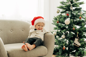 A cheerful boy in a Santa Claus hat is sitting under a Christmas tree in an armchair and enjoying the holidays