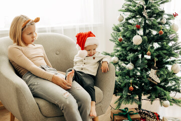 Charming children are sitting in a chair in a room decorated for Christmas and New Year. A boy in a Santa Claus hat and his sister in tiger ears. The concept of Christmas and New Year - Powered by Adobe