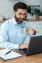 Businessman in elegant shirt front of laptop looking at screen drinking coffee
