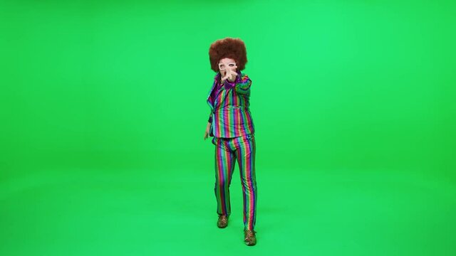 Retro dude in a funny wig and multicolored suit is dancing a funny dance on a green background, festive mood, a retro disco man is having fun at a new year's party.