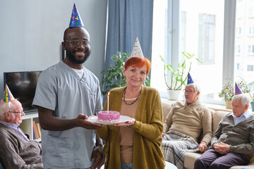 Portrait of African doctor smiling at camera while celebrating senior woman with birthday and giving a cake for her