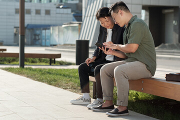 Young businessman in casualwear showing his female colleague online video in his smartphone while both sitting on bench