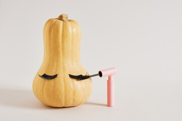 mascara in pink packaging and pumpkin with false eyelashes on gray background copy space