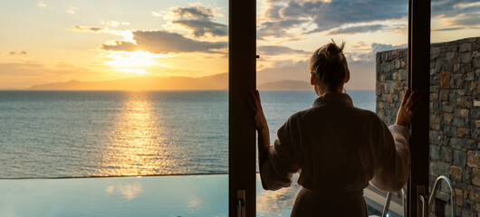 woman in bathrobe standing between sliding glass doors and enjoying sunrise over the sea in luxury villa