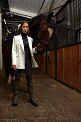 Fototapeta premium Portrait of smiling female jockey standing by horse in stable