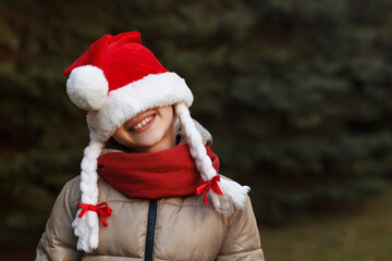 Christmas child in santa hat with pigtails in new year tree background. Smiling kid girl is happy.