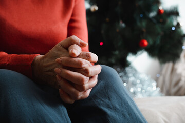 Lonely senior woman sitting at home in Christmas celebration. Close-up of an elderly woman's hand against background of decorated Christmas tree. Loneliness, sad holiday concept