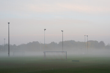 Empty and deserted soccer field in suburban area with goals and flood light poles on grey overcast day with fog and mist
