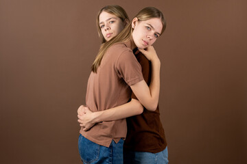 Affectionate teenage twins in blue jeans and brown shirts looking at you while giving each other hug in front of camera