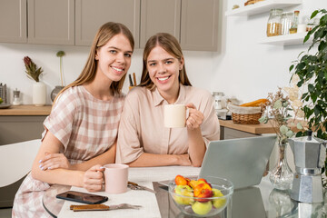 Cute and cheerful twin girls with drinks sitting by kitchen table in front of camera while enjoying weekend