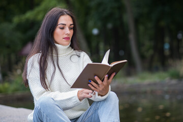 Obraz premium A young and attractive Caucasian brunette girl sitting and reading a book in a park near the river. The student is studying. The concept of education and knowledge.
