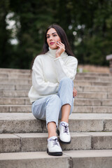 Fototapeta premium A beautiful and attractive Caucasian brunette girl in a white sweater sitting on the steps in the park.