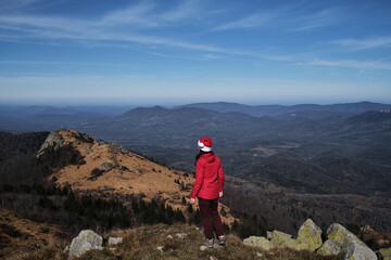 Woman in red Santa hat celebrates Christmas and New Year on top of mountain without snow in warm country. Stand with back turned and enjoy scenery and wildlife views. Tourist travels in red jacket.