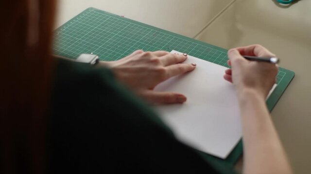 Close-up view from back of unrecognizable young woman making mark with pencil on white paper lying on green rubber cutting mat sitting at desk, selective focus. Tracking shot in slow motion.
