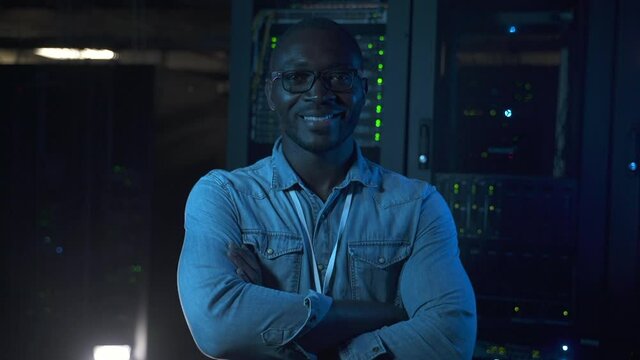 Young male engineer posing for camera and smiling during day or night at data office spbas. Front view of american african man looks ahead with smile and poses with crossed arms, stands in dark
