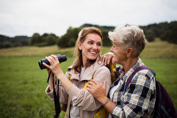Happy senior mother hiker embracing with adult daughter holding binoculars outdoors in nature