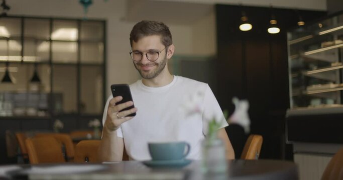 Handsome man in glasses wearing a white T-shirt uses a smartphone and smiles at coffee shop. Cheerful man surfing the Internet by mobile phone while spending free time in a cozy cafe. 