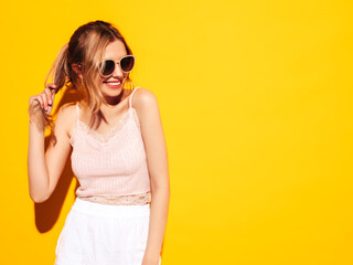 Portrait of young beautiful smiling female in trendy summer clothes. Sexy carefree woman posing near yellow wall in studio. Positive model having fun indoors. Cheerful and happy. In sunglasses
