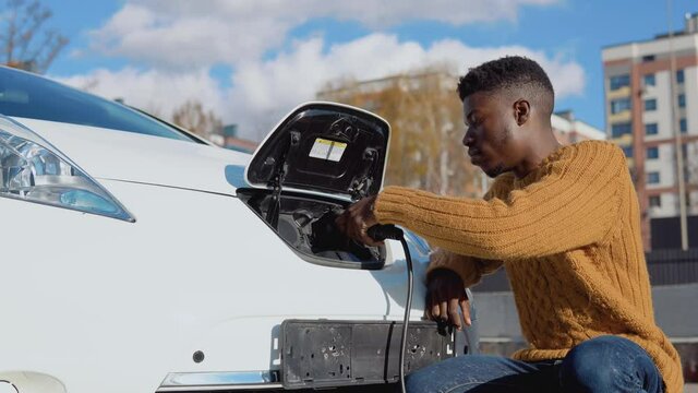 African american male driver connects electric car to power system to charge car battery