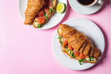 Croissant sandwich with cream cheese, salmon and arugula on white plate, pink background. Healthy breakfast concept.