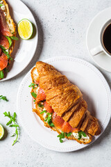 Croissant sandwich with cream cheese, salmon and arugula on white plate, gray background. Healthy breakfast concept.
