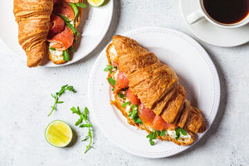 Croissant sandwich with cream cheese, salmon and arugula on white plate, gray background. Healthy breakfast concept.