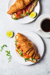 Croissant sandwich with cream cheese, salmon and arugula on white plate, gray background. Healthy breakfast concept.
