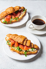 Croissant sandwich with cream cheese, salmon and arugula on white plate, gray background. Healthy breakfast concept.