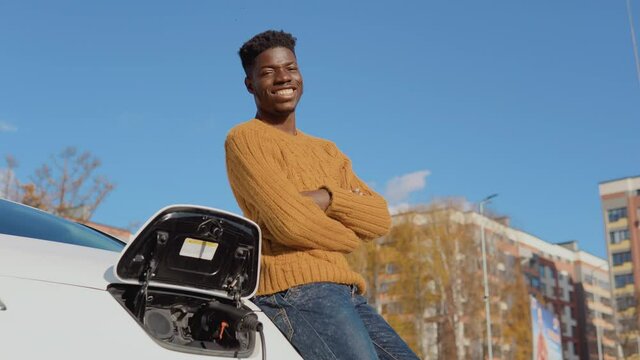 A dark-skinned driver is standing near a white electric car while charging the battery