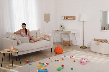 Happy young mother with cup of coffee and laptop resting in messy living room