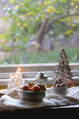 Silver bowl filled with fresh tangerines, various Christmas decorations, lit candles and soft blanket. Hygge at home. Selective focus.