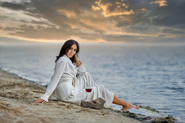 A beautiful girl in a white suit with a glass of red wine sits on the beach at sunset. High quality photo