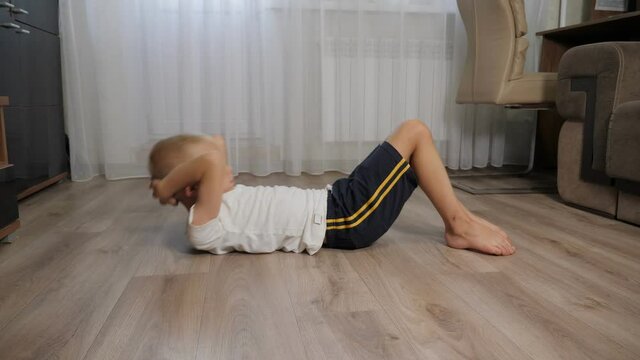 A little eight-year-old boy in shorts and a T-shirt shakes his abs lying on the floor at home. Training at home during quarantine.