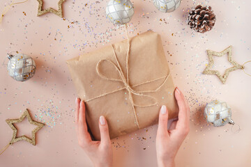 Female hands with natural manicure holding present in kraft paper with jute on soft beige background. Xmas composition. Flat lay. Happy holidays, New Year celebration and giving love concept. 
