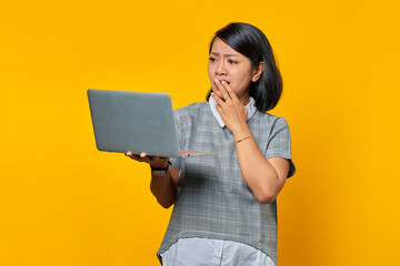Shocked Asian woman looking at laptop with hand covering mouth over yellow background