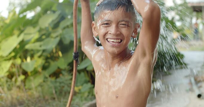 Slow motion scene of an Asian boy who is the farmer's son in a rural area uses a hose to pour water over his head to take a shower outside the house with a laugh and happiness.