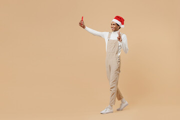 Full body young african man in red Christmas hat do selfie shot mobile cell phone post photo on social network isolated on plain pastel beige background studio Happy New Year 2022 celebration concept