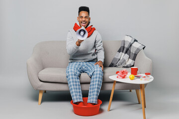 Young african man in sweater scarf sit on sofa hold scream in megaphone announces discounts sale...