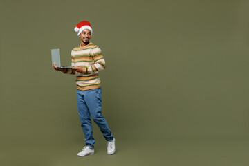 Full body young african man in knitted sweater red Christmas Santa Claus hat hold use work laptop pc computer isolated on plain green khaki background studio Happy New Year 2022 celebration concept