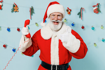 Old Santa Claus man 50s wear Christmas hat red suit clothes talk speak on handset phone show thumb up isolated on plain blue background studio. Happy New Year 2022 celebration merry ho x-mas concept.