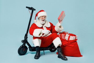 Full body old Santa Claus man in Christmas hat red suit clothes sit on scooter with gifts bag hold gift box isolated on plain blue background studio Happy New Year 2022 merry ho x-mas holiday concept