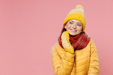 Pensive wistful happy young woman 20s years old wears yellow jacket hat mittens put hand prop up on chin lost in thought and conjectures isolated on plain pastel light pink background studio portrait.